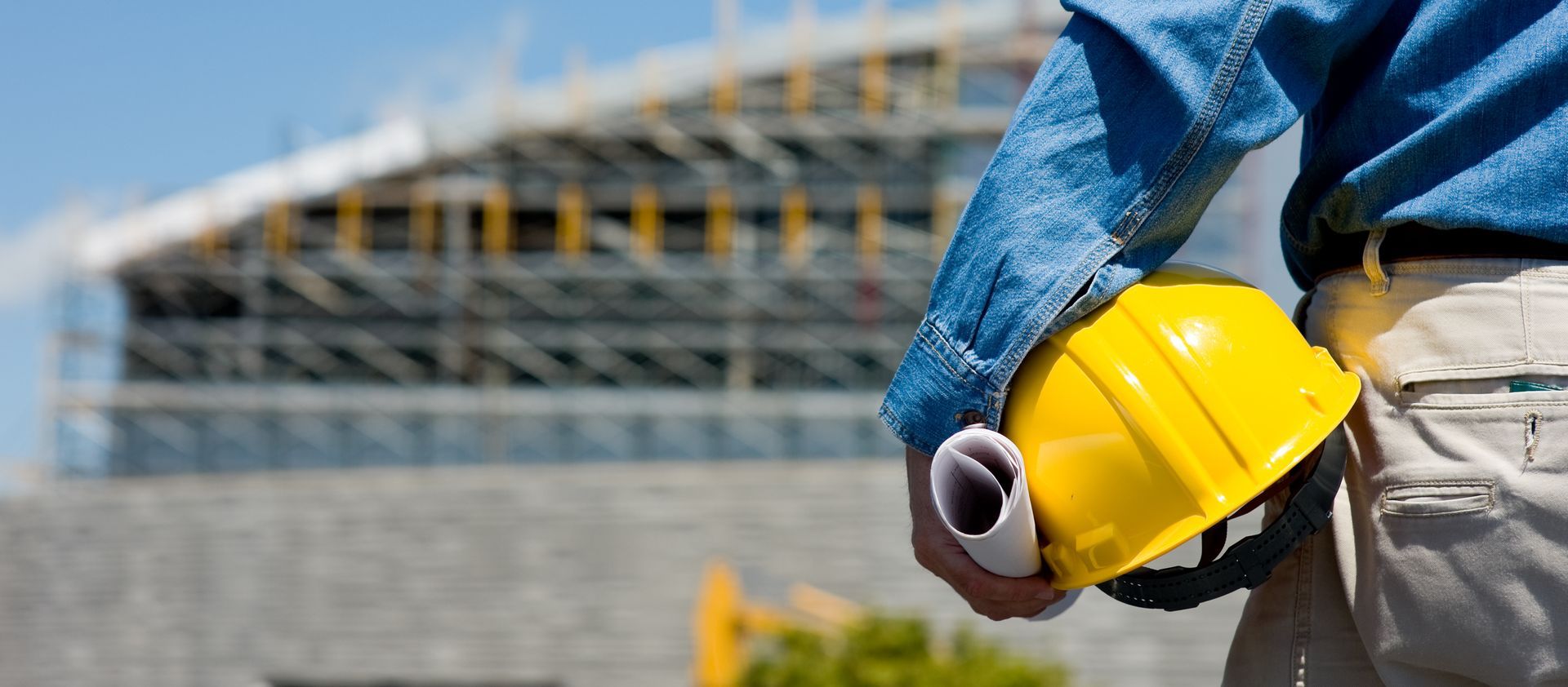 Construction worker holding a yellow hard hat and rolled blueprints, with a building under construction in the background. Construction worker holding a yellow hard hat and rolled blueprints, with a building under construction in the background.