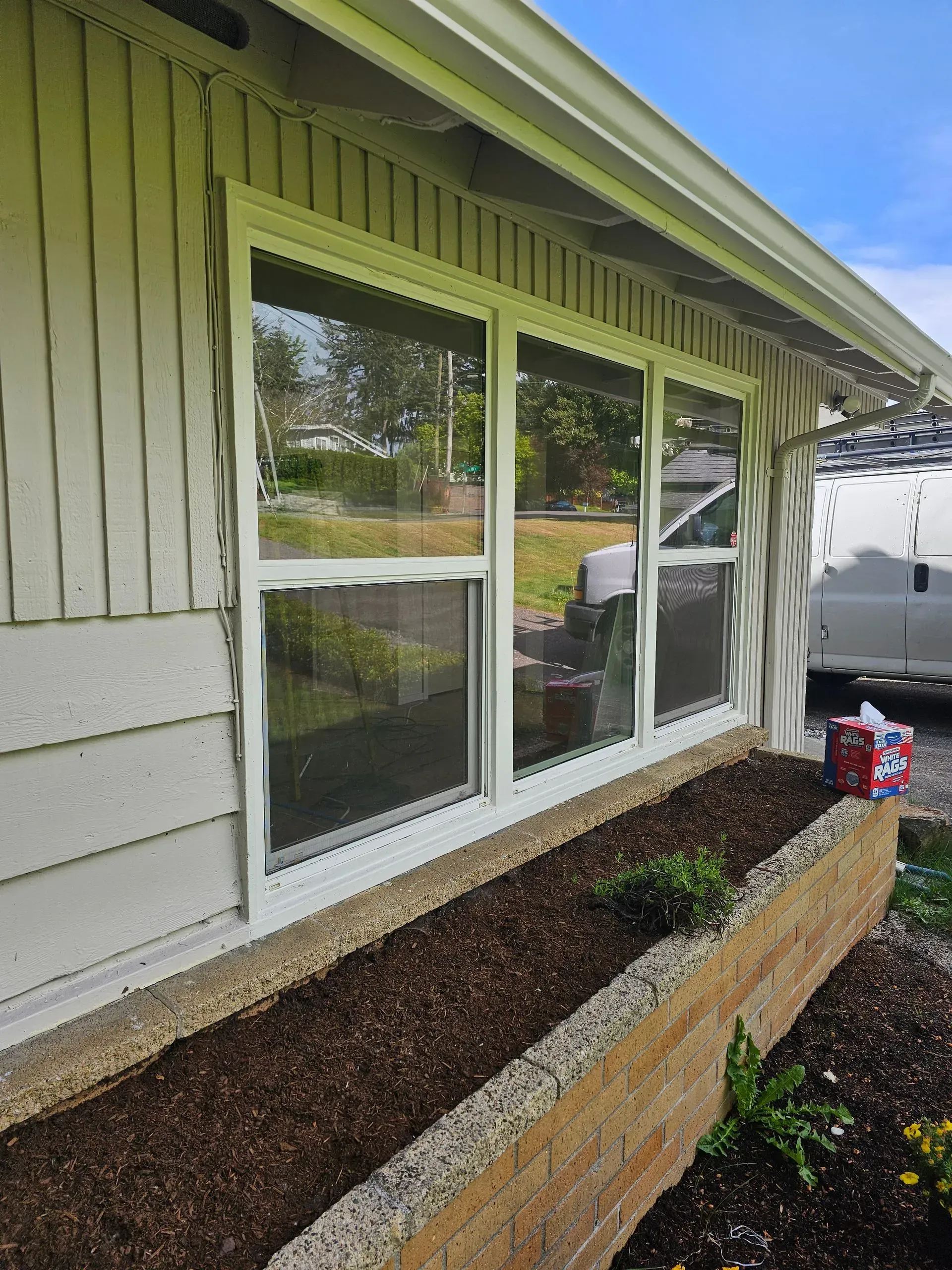 White, vertical-siding house exterior with three windows above a brick-edged planter bed filled with dark mulch.