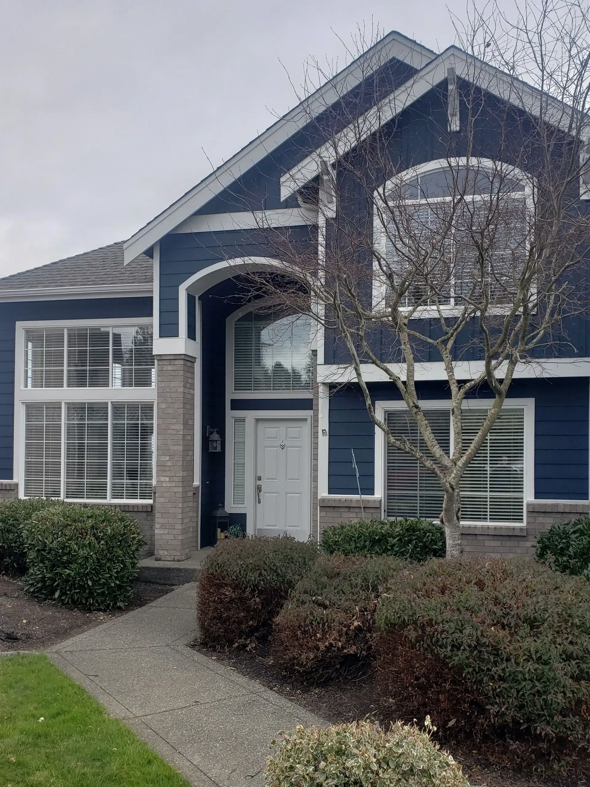 Two-story blue house with white trim, a stone-accented arched entryway, large windows, and manicured shrubs in the yard.