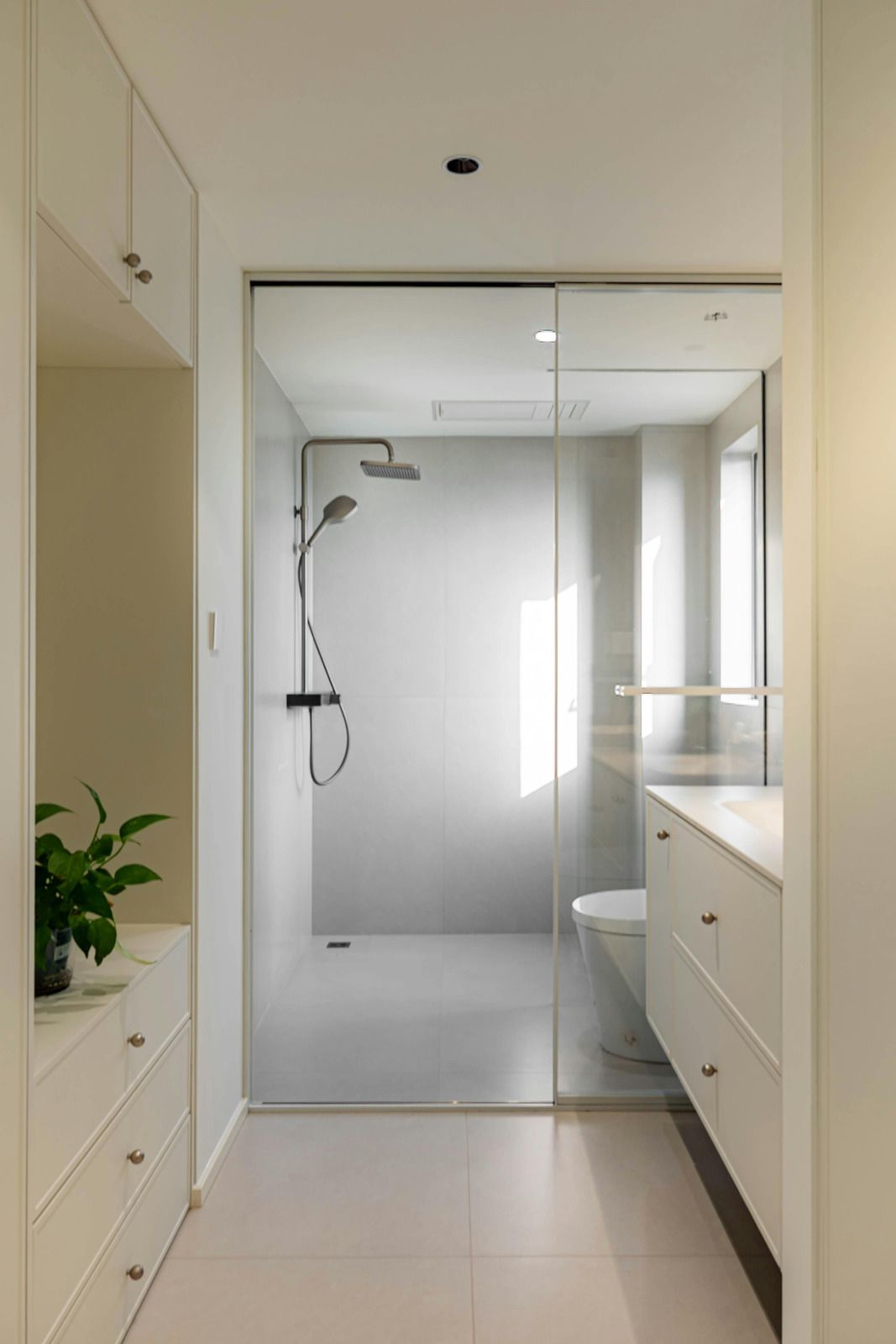 A modern, minimalist bathroom with white cabinets, a glass shower stall, and light-toned tiled flooring.