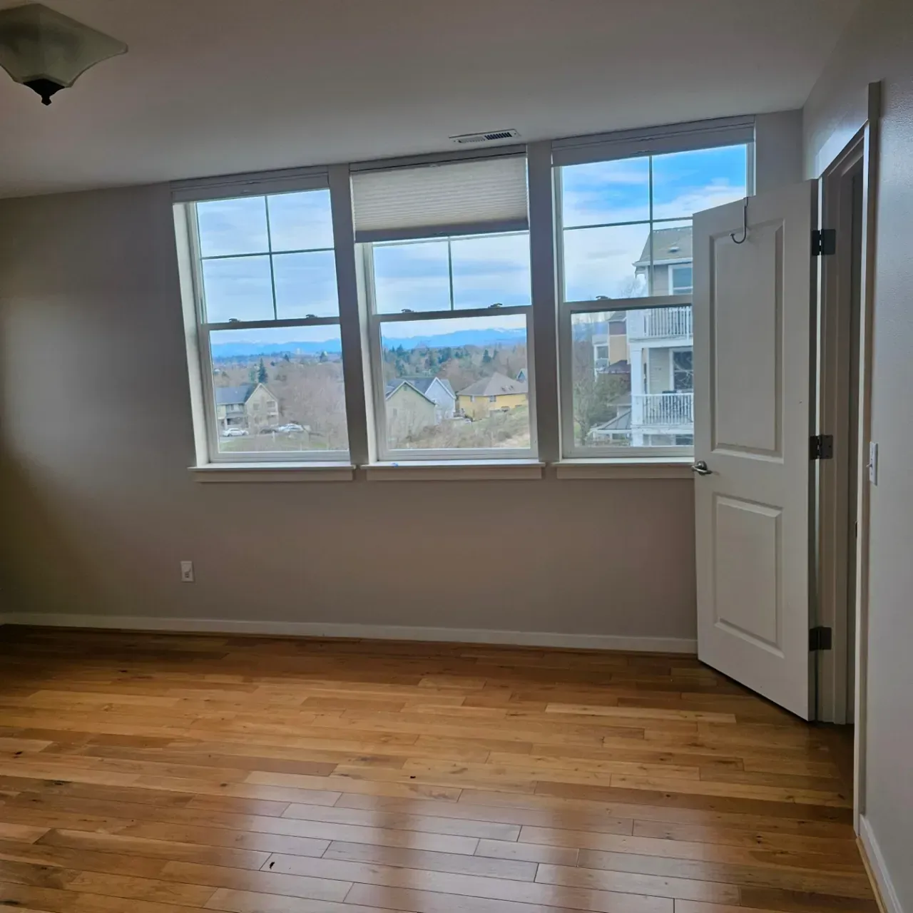 An empty room with hardwood floors, neutral-toned walls, a ceiling light, and a large window looking out onto a landscape.