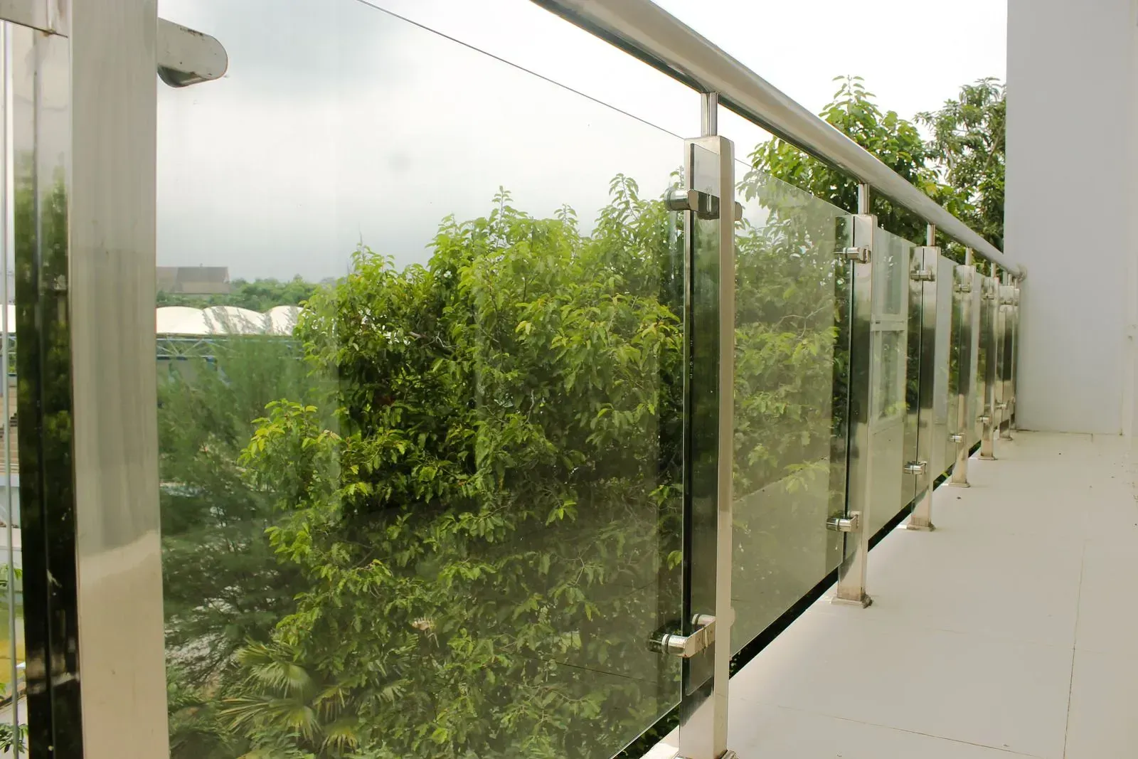 A balcony railing made of glass panels supported by polished metal posts, overlooking lush green trees.