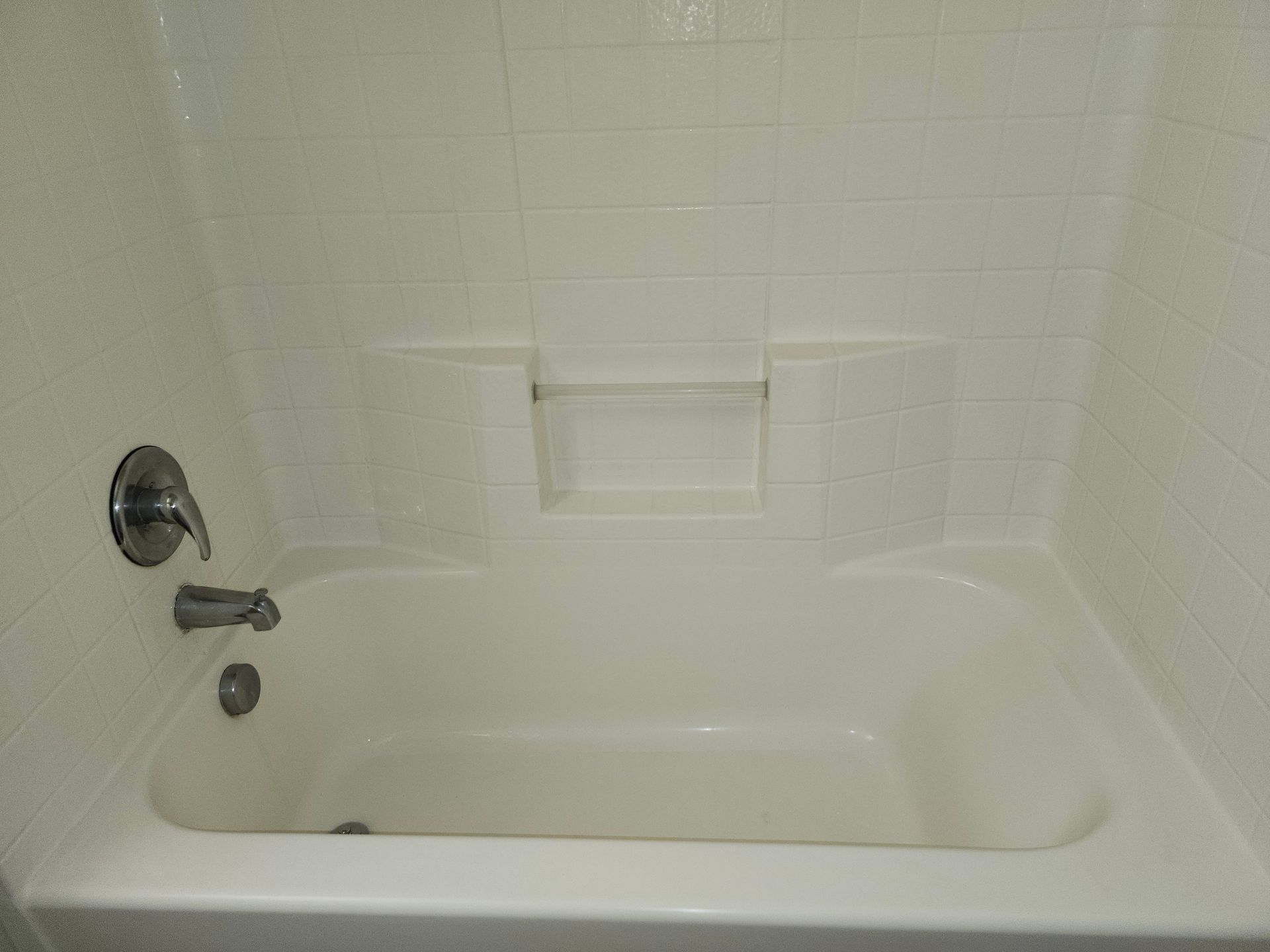 White bathtub with white tiled walls, silver faucet, and built-in shelf.