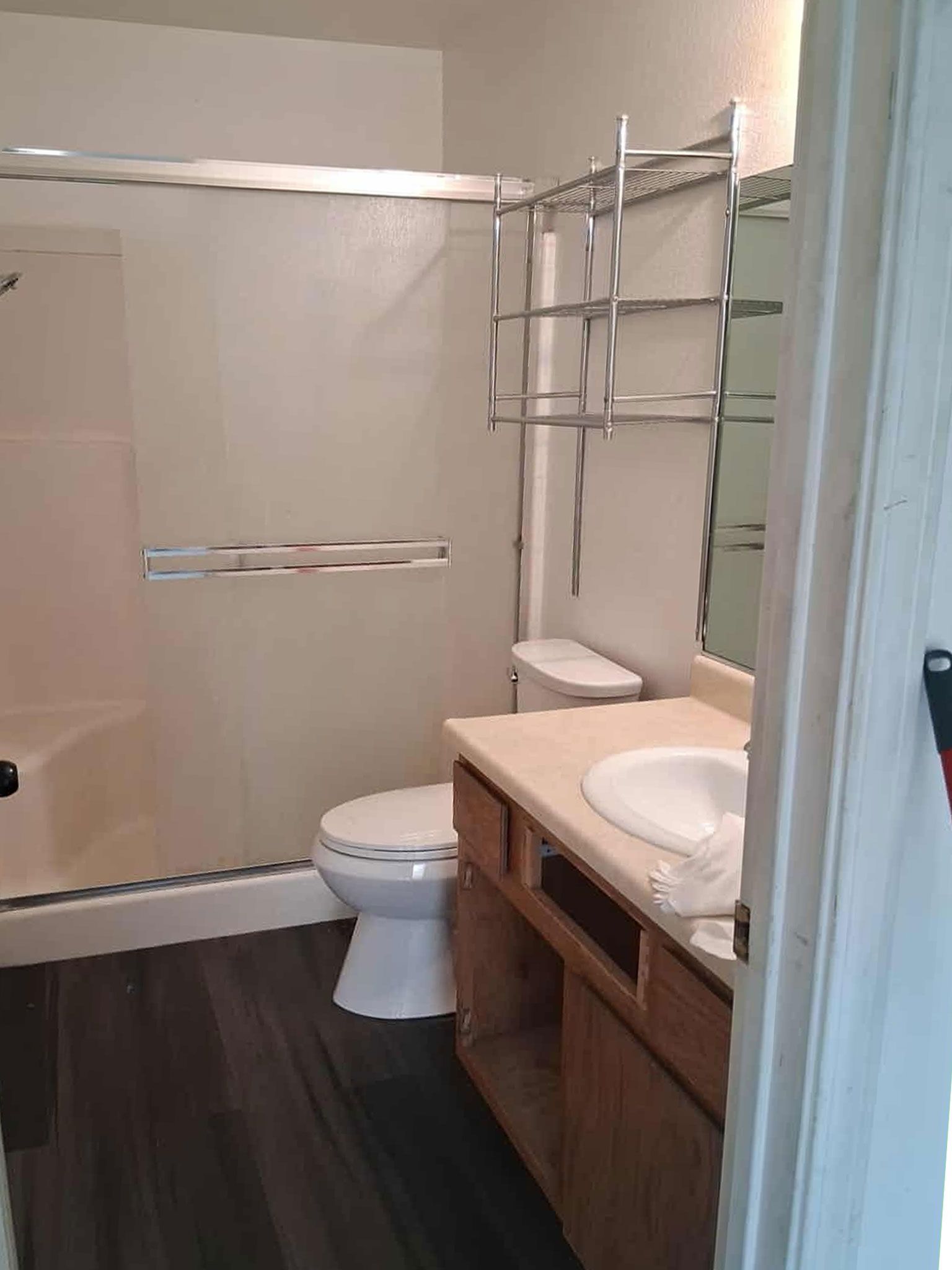 Bathroom with shower, toilet, vanity, and shelving. Dark floor, white fixtures, and tan cabinets.