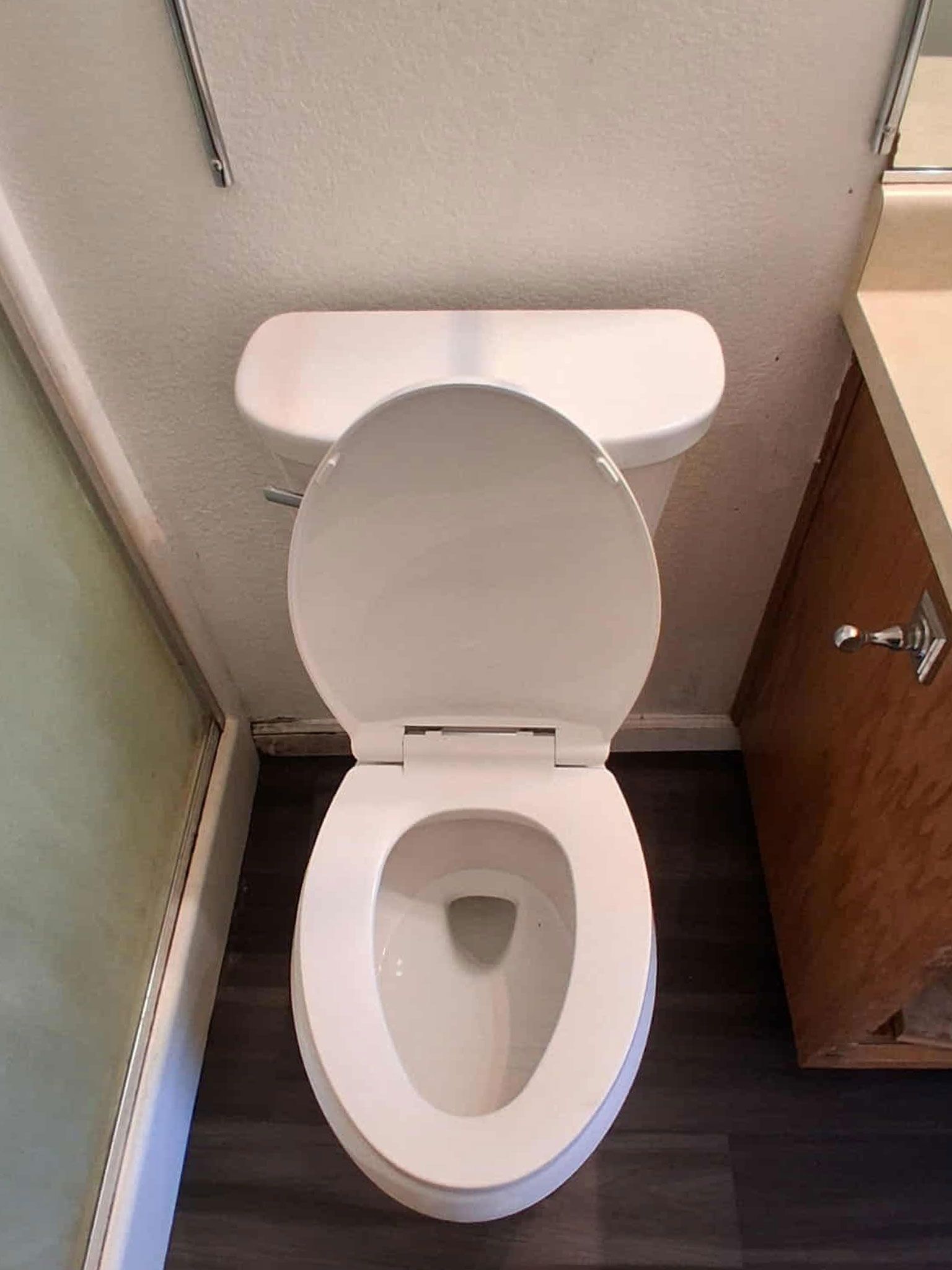 White toilet with open seat in a bathroom. Dark wood-look floor and brown vanity are visible.