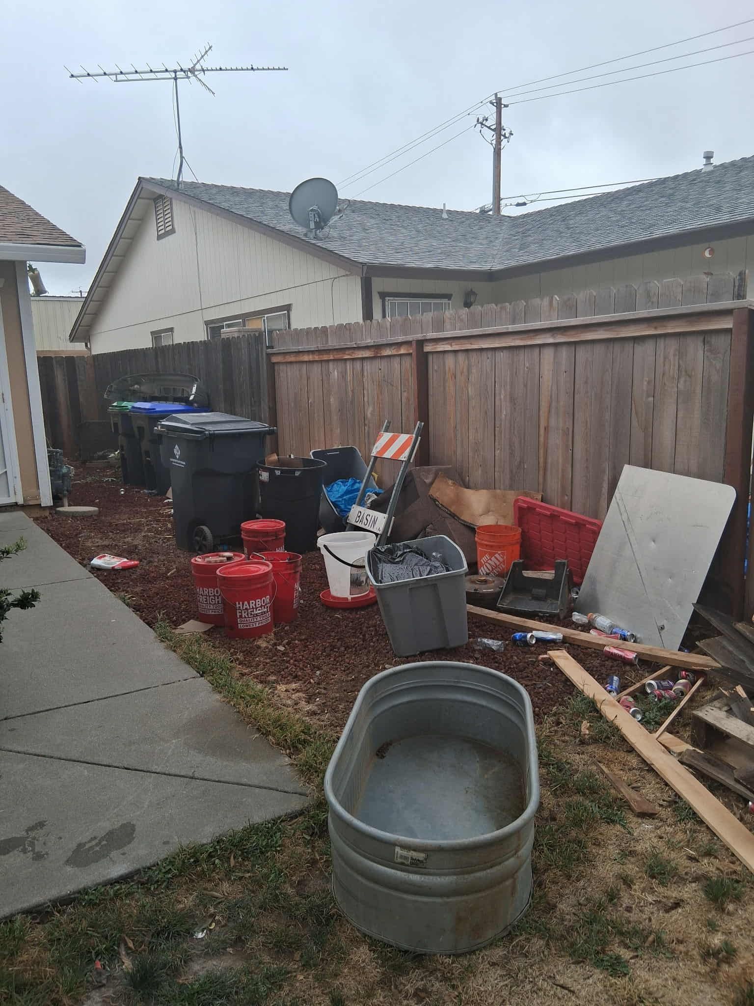 Backyard with trash cans, debris, and a metal trough near a fence and house.