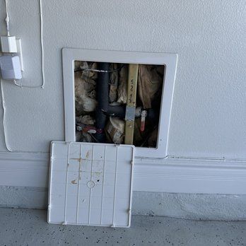 Square opening in white wall, revealing plumbing and insulation. White access panel is open and on the floor.