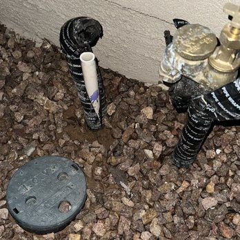 Close-up of irrigation system components, including pipes, valves, and a round access cover, surrounded by gravel.