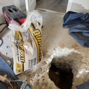 Bag of Quikrete sand sits near a hole in a concrete floor, near a doorway. A toolbox and tarp are also in the room.