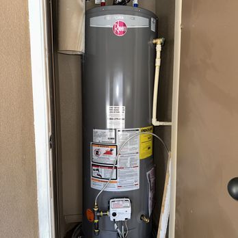 A tall, gray Rheem water heater installed in a narrow closet. White pipes and controls are visible.
