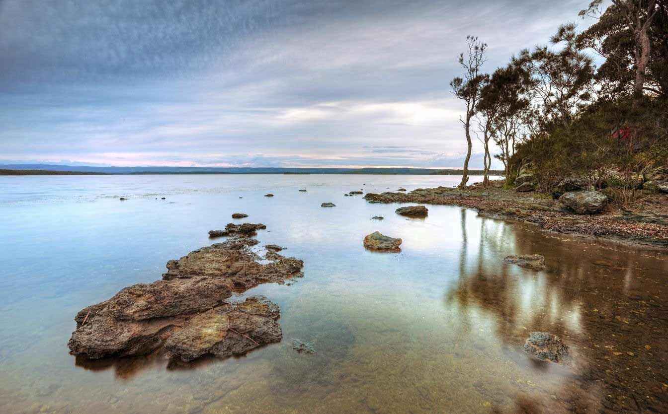 A Large Body Of Water With Rocks In The Foreground And Trees In The Background — Sal's Excavations In St. Georges Basin, NSW