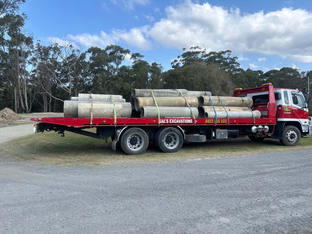 A Red Tow Truck Carrying Concrete Pipes On A Flatbed Trailer — Sal's Excavations In Falls Creek, NSW