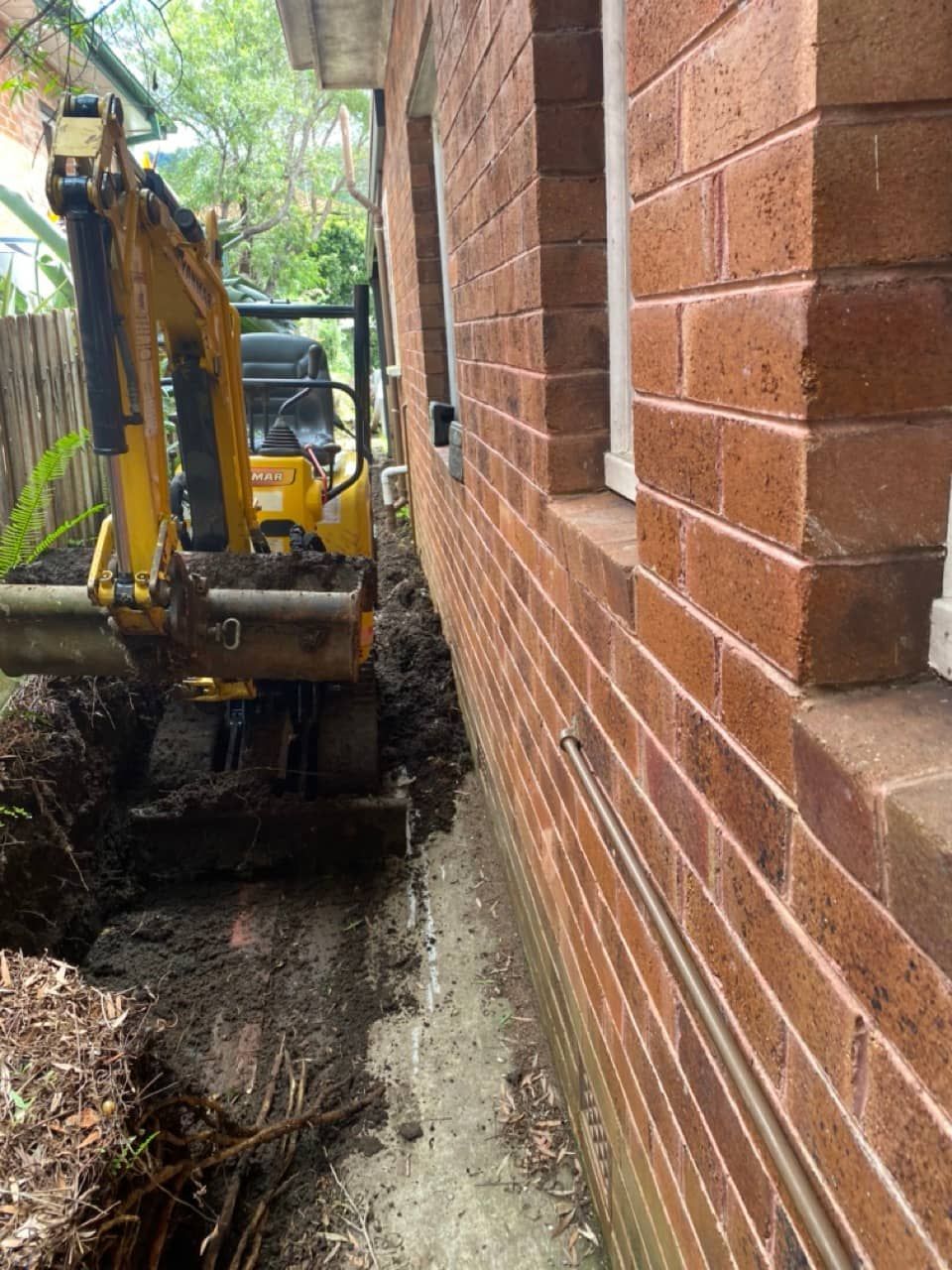 A Yellow Excavator Digging A Hole In The Side Of A Brick House — Sal's Excavations In Falls Creek, NSW