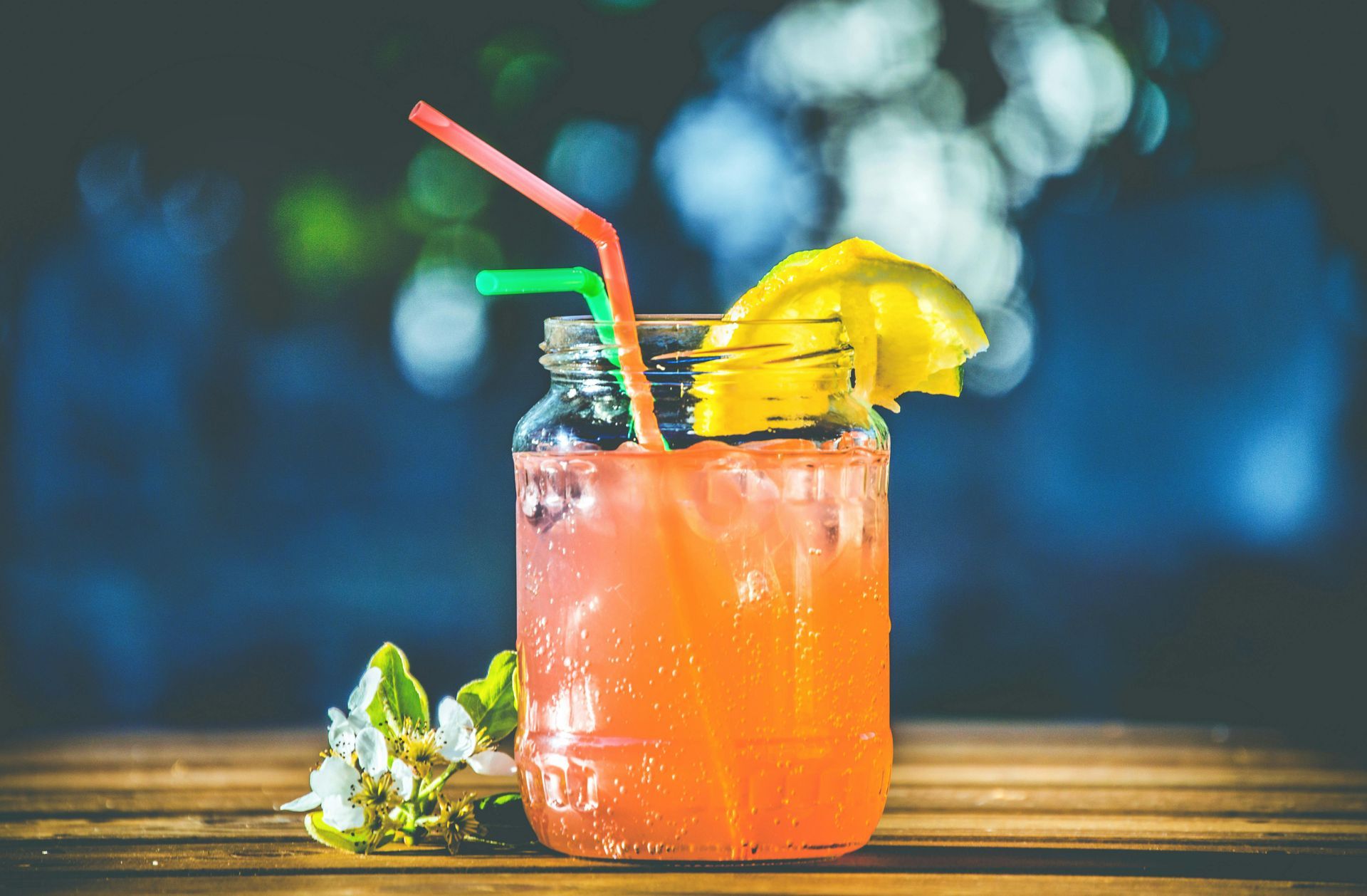 Glass of orange-pink drink with lemon garnish and straws on a wooden table. Blurred blue background.