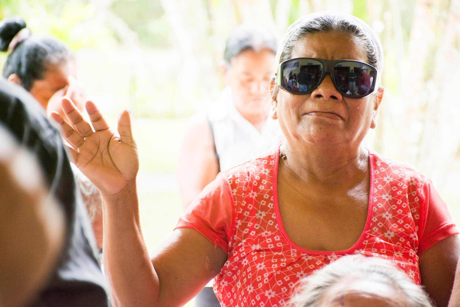 A woman wearing sunglasses and a red shirt is waving her hand.