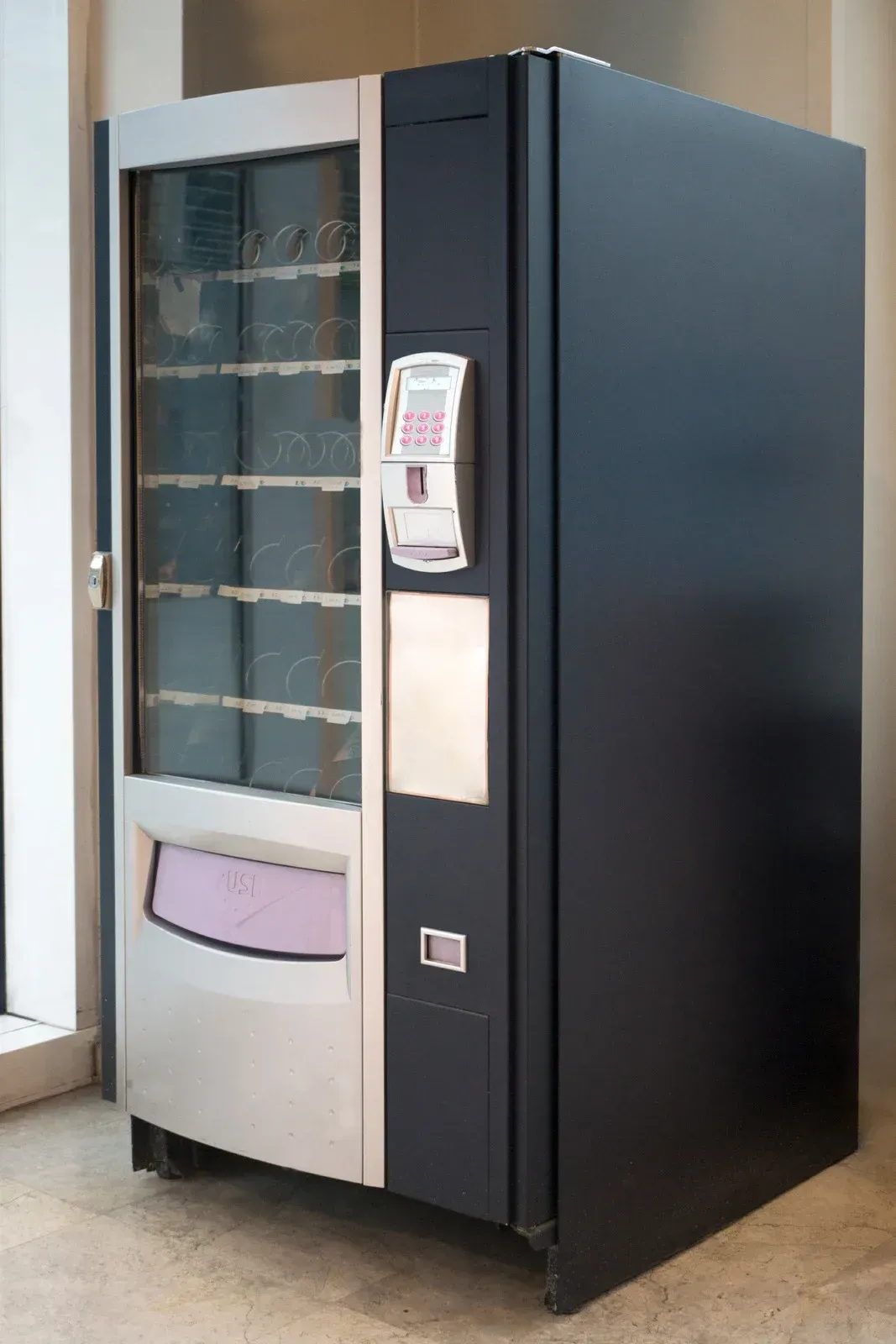 A vending machine is sitting in a room next to a window.