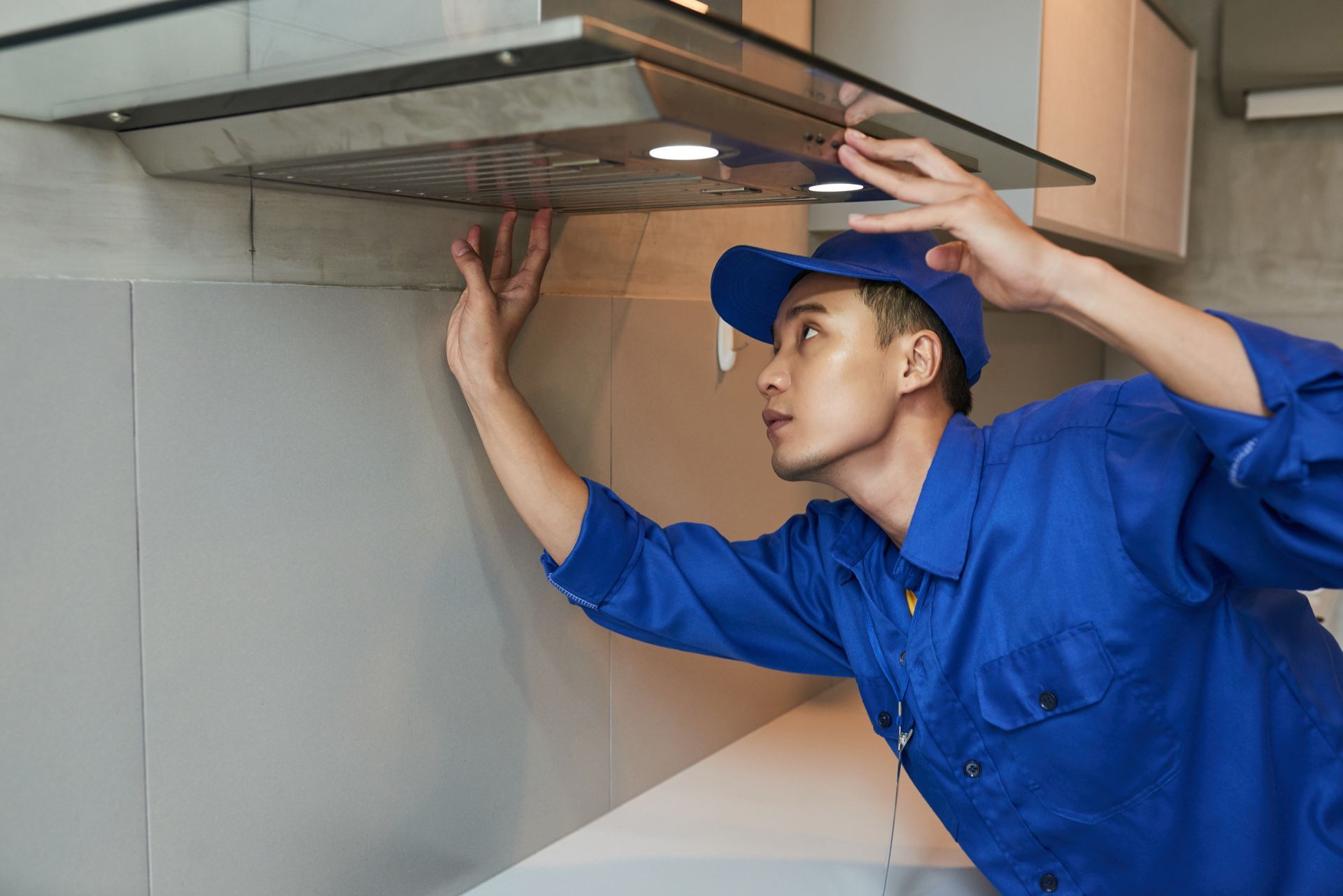 A man in a blue shirt and hat is fixing a hood in a kitchen.