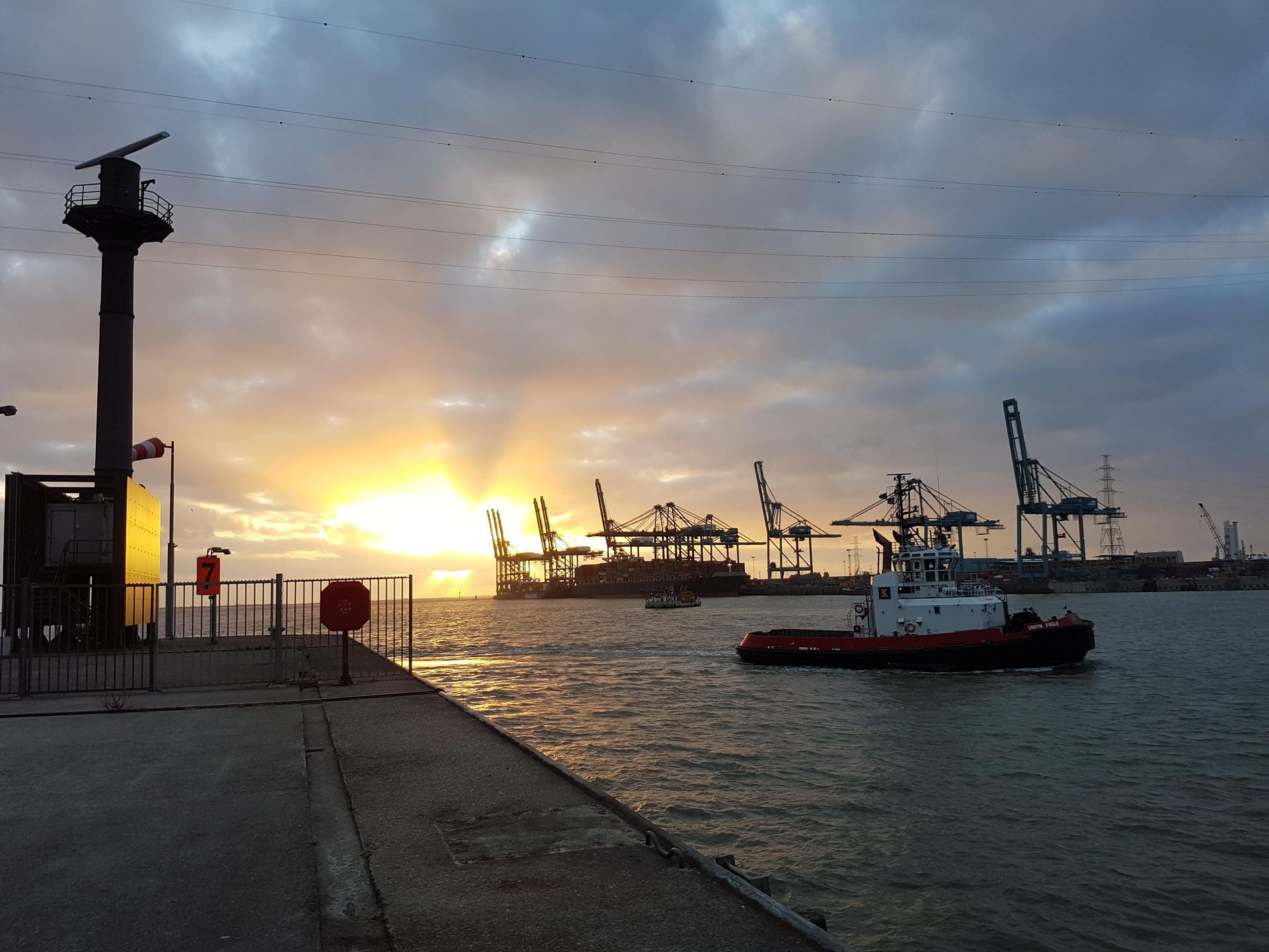 Sunrise over a harbor with ships, cranes, and a lighthouse.