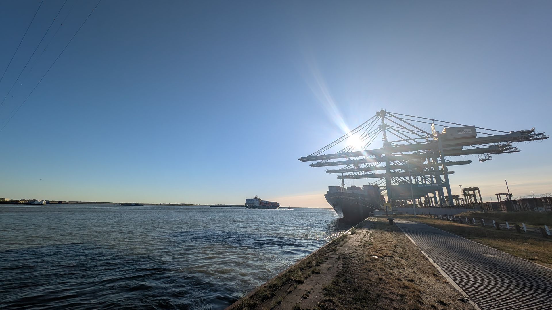 Sun shining behind a cargo ship at port, with large cranes and a blue sky.