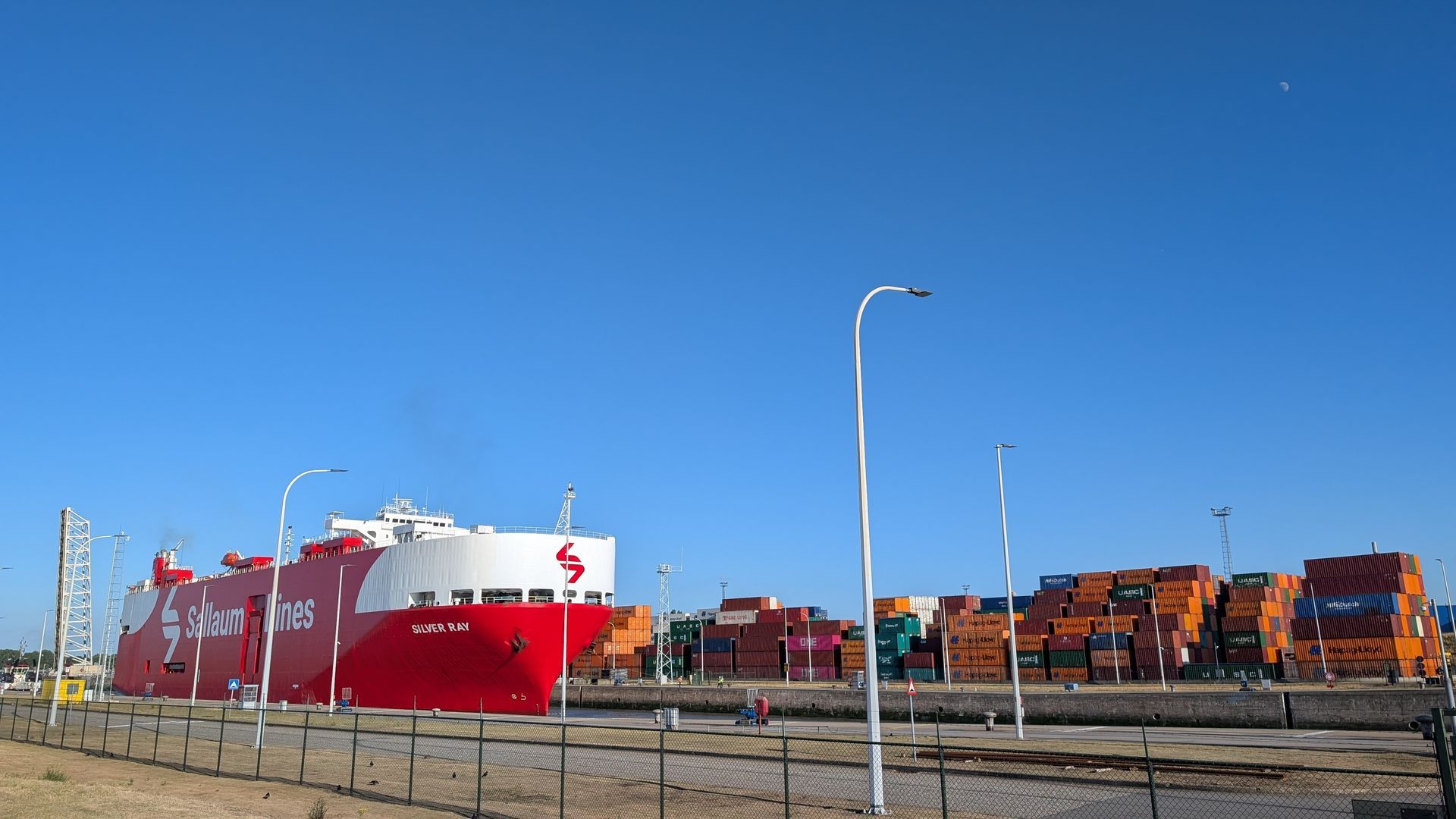 Een groot rood vrachtschip in een haven met op de achtergrond gestapelde containers tegen een blauwe lucht.