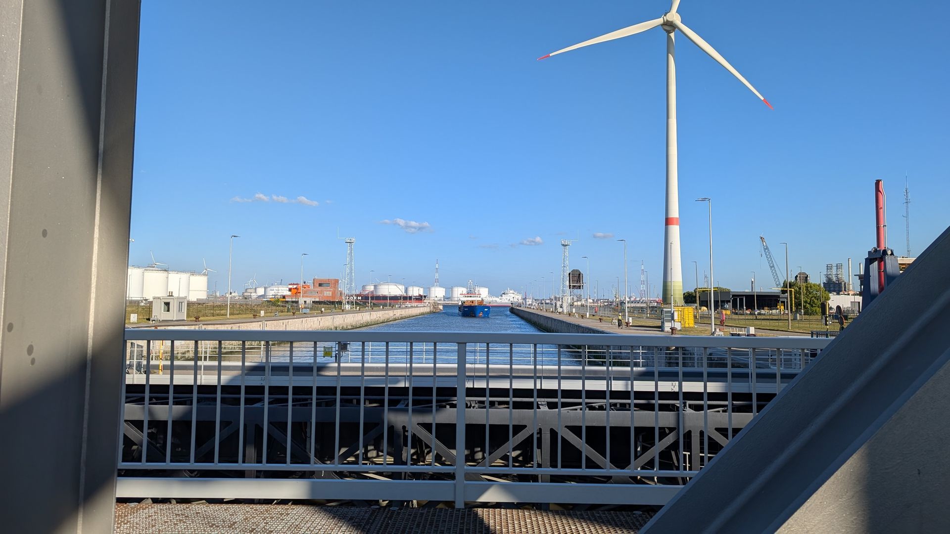 A wind turbine stands next to a canal, viewed from a bridge under a clear blue sky.