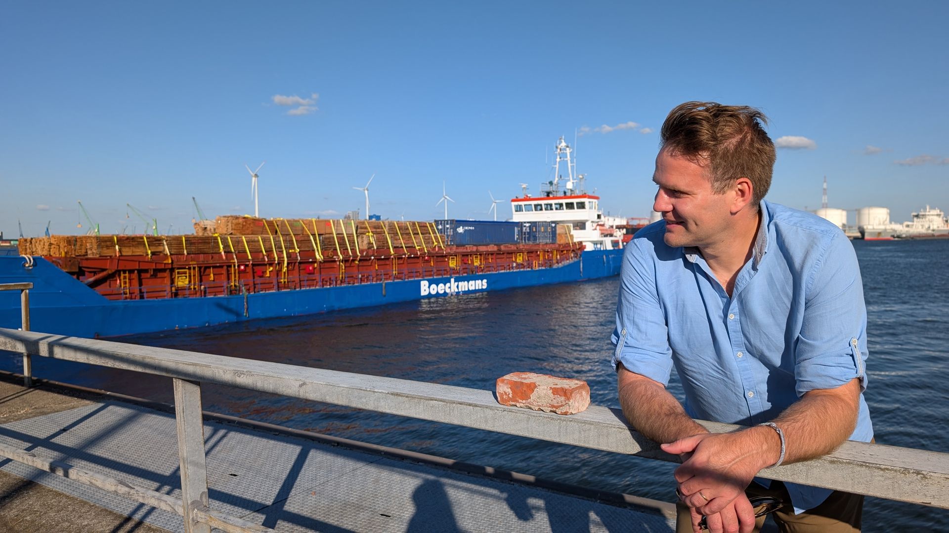 Een man leunt tegen de reling, glimlachend, met een blauw schip en een haven op de achtergrond. Blauwe lucht.