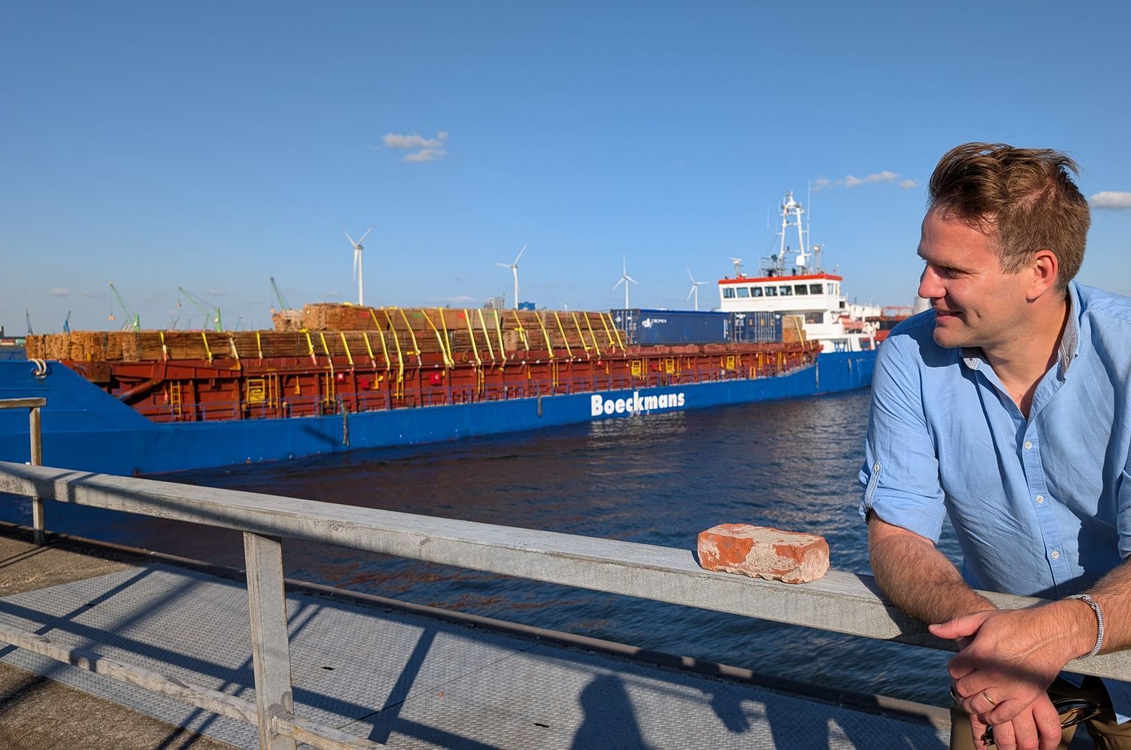 Man smiles, leaning on railing, looks toward blue cargo ship in harbor under a clear sky.