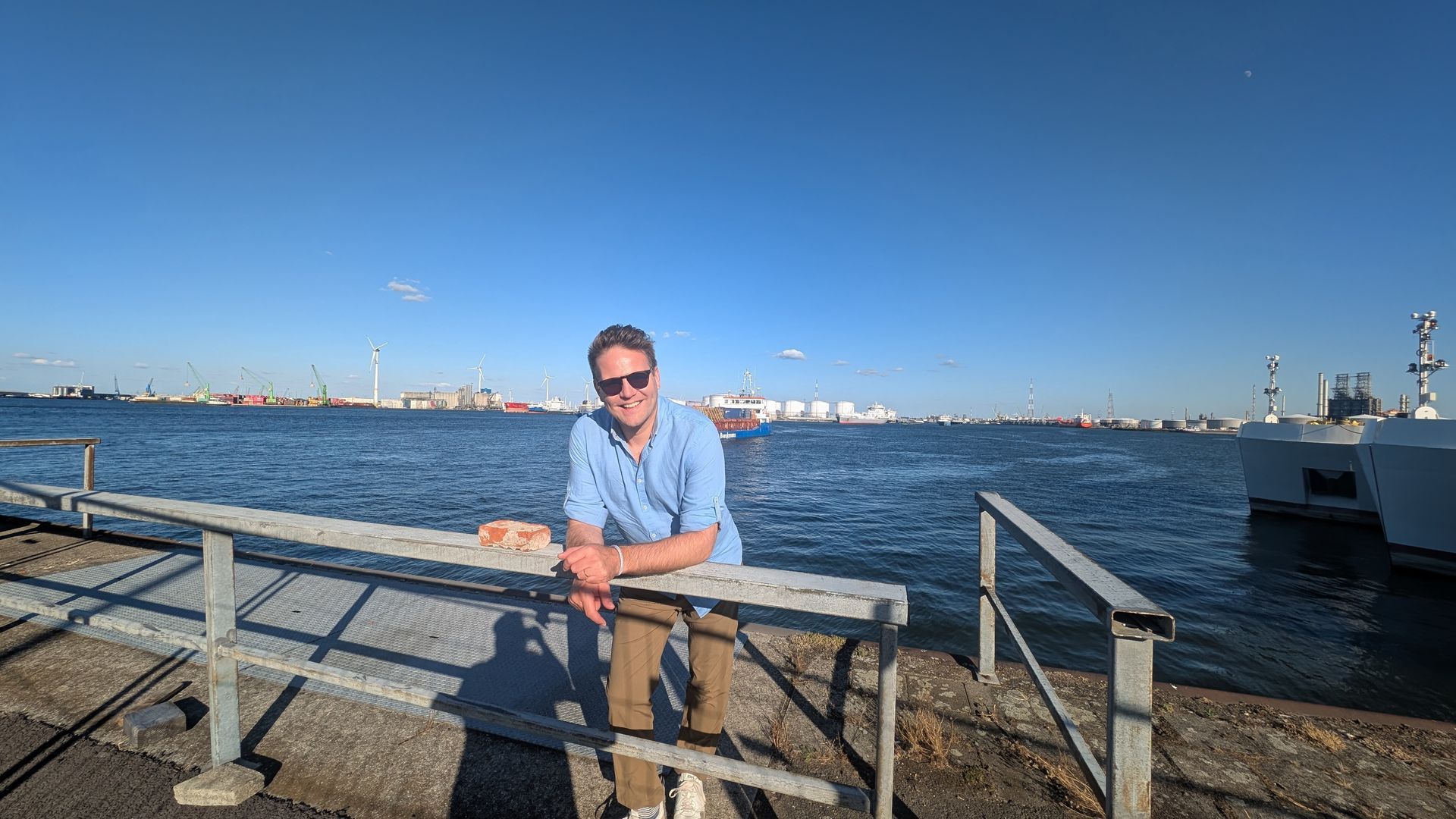 Man in sunglasses leans on a railing at a harbor, facing the water with blue sky overhead.