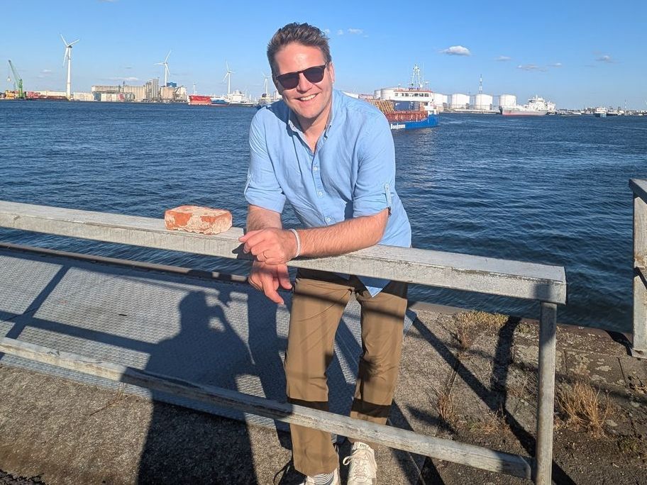 Man in sunglasses leaning on a railing by water, smiling. Port in background.