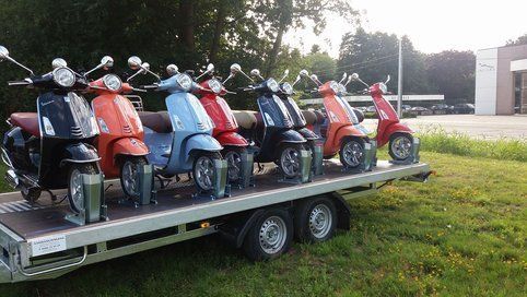Op een zonnige dag staan scooters in verschillende kleuren op een aanhanger op een grasveld opgesteld.