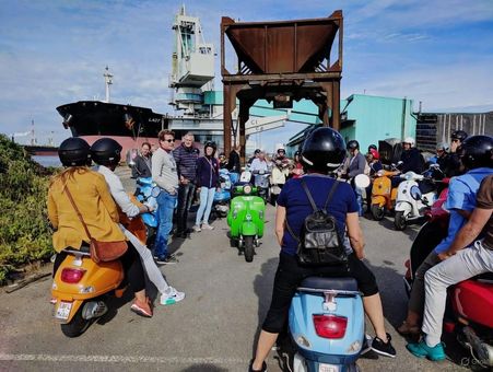 Group on scooters in front of a structure and ship. Several colorful scooters with riders are in formation.