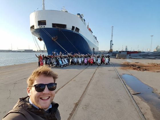 Man taking selfie with group of people on scooters in front of a large cargo ship at a port.