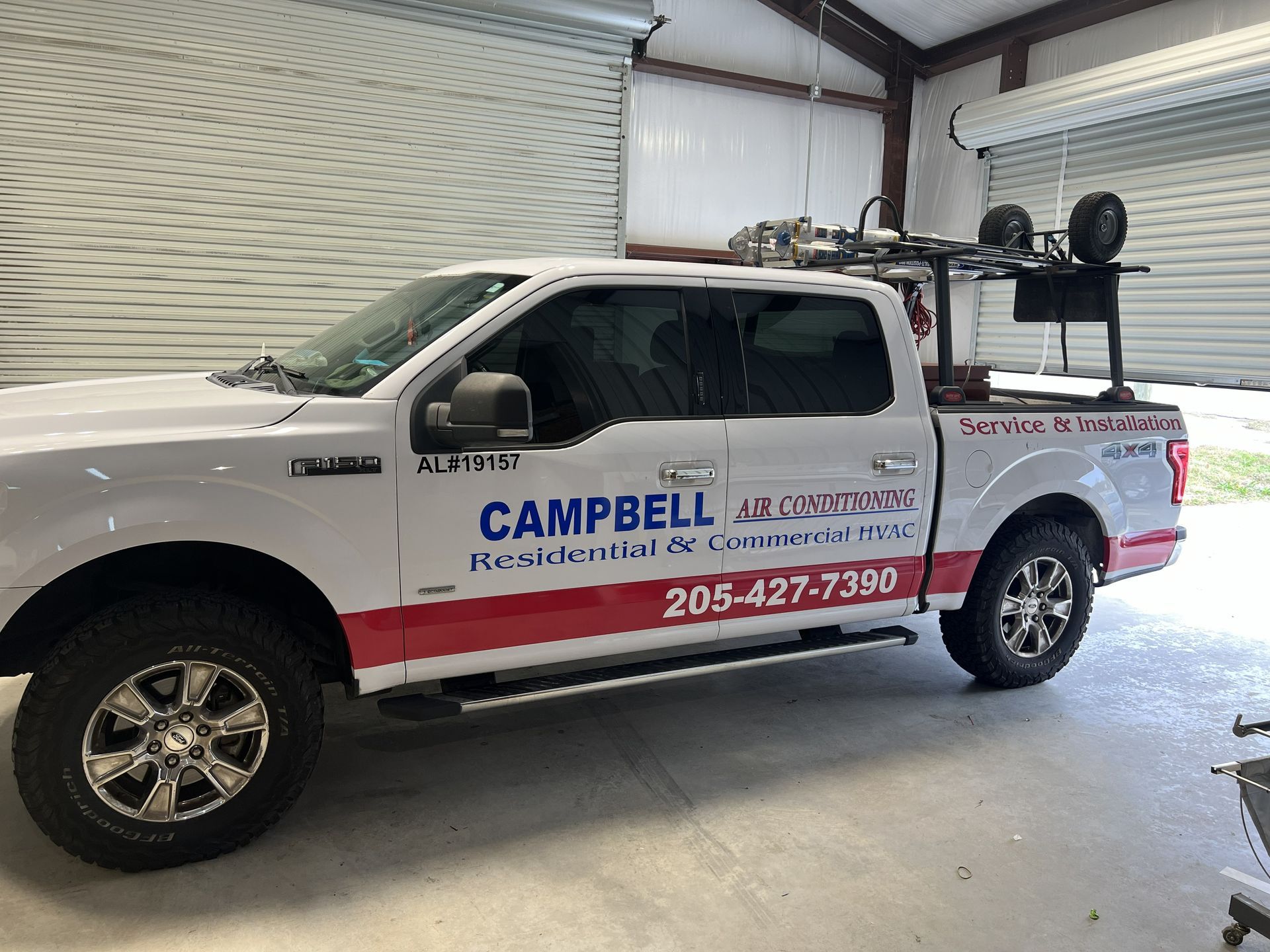 White Campbell Landscaping truck parked in a garage. Red stripe, equipment rack. Phone number visible.