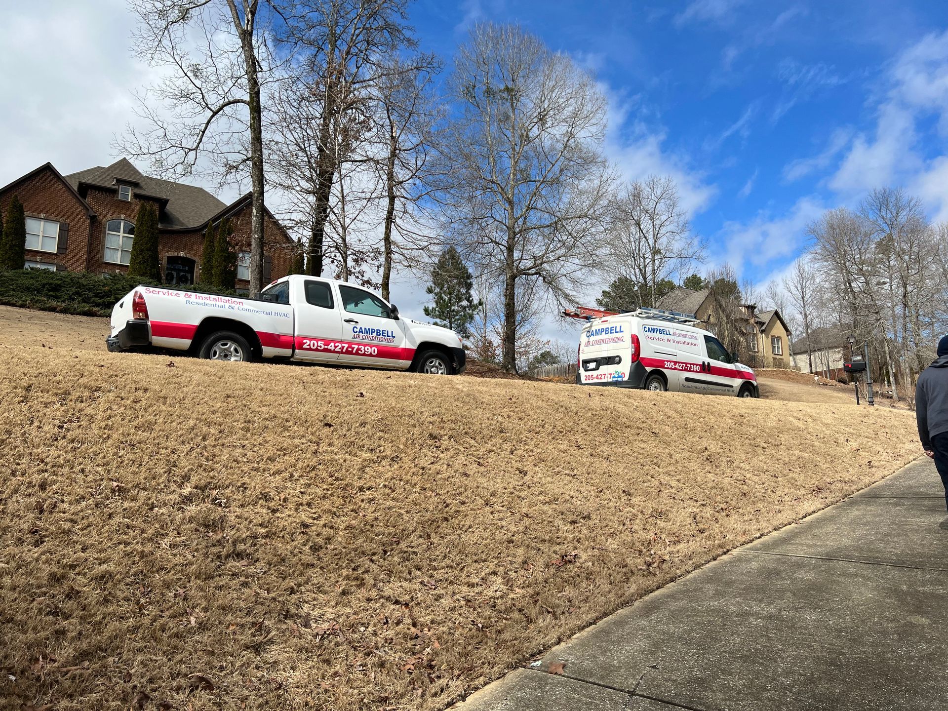 Two service vehicles parked on a grassy slope, in front of a brick house, under a cloudy sky.