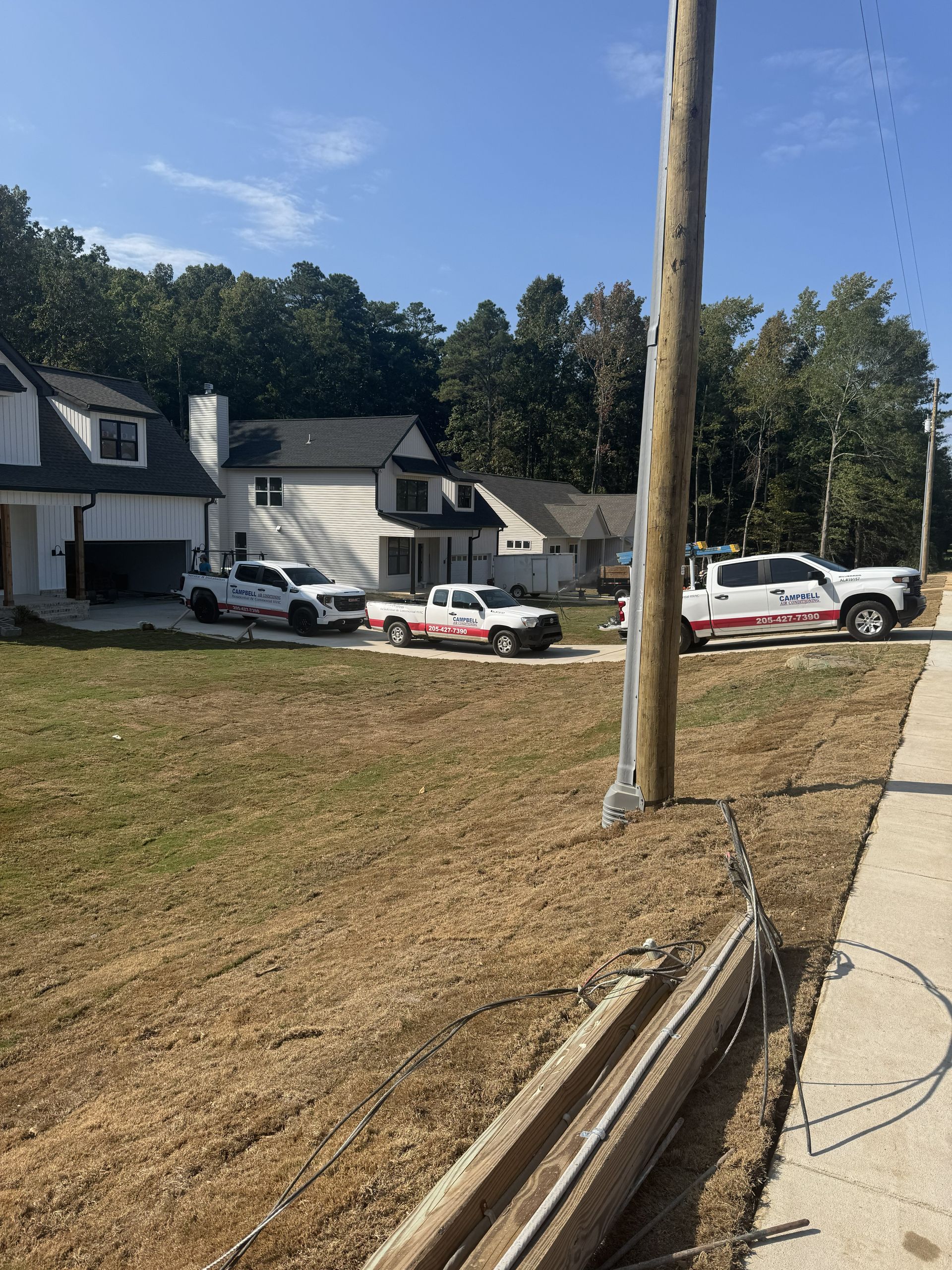 Three white work trucks parked in front of new homes under construction. Grassy lot with trees and a utility pole.