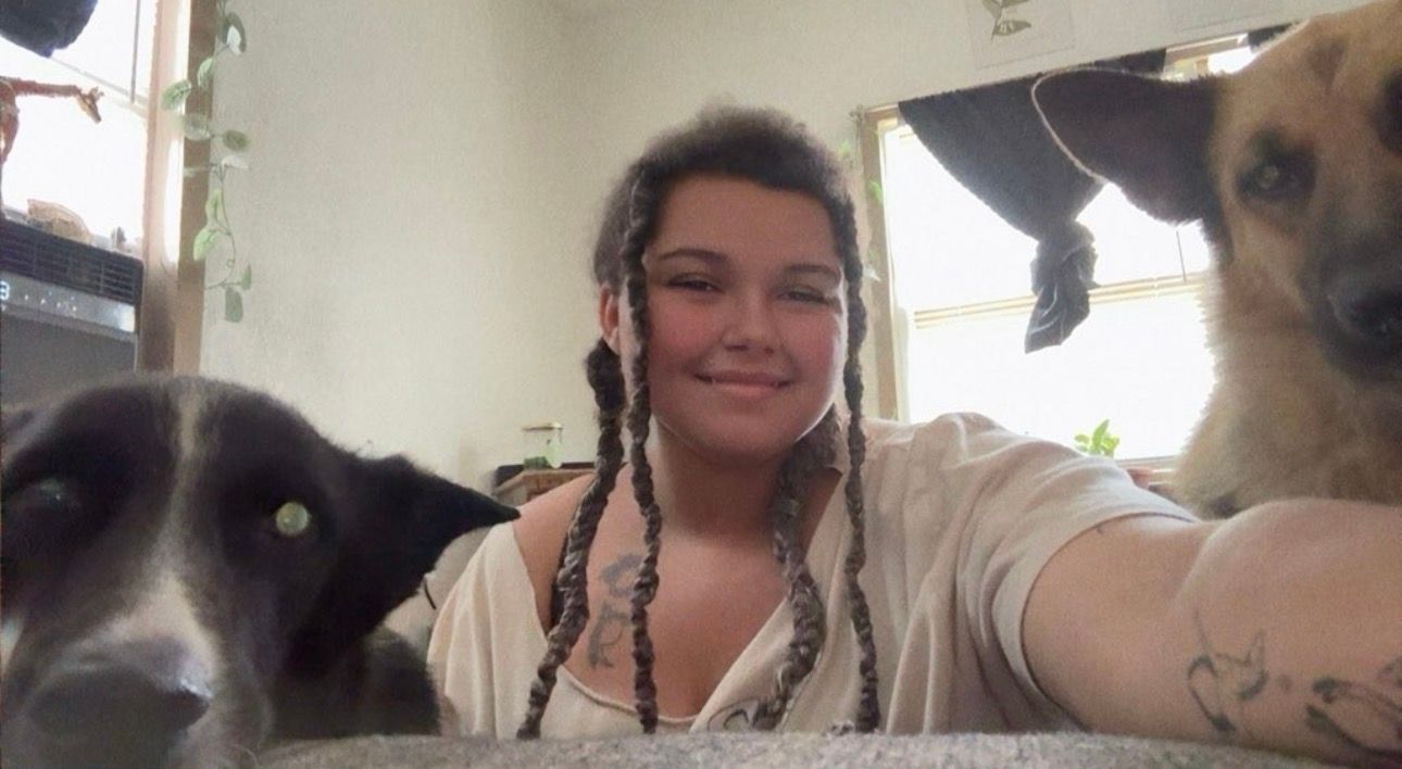 Young person smiling between two dogs indoors, with one black dog close left and one tan dog close right.