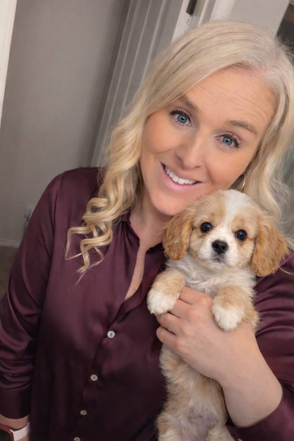Woman with blonde hair smiling, holding a small beige and white dog.