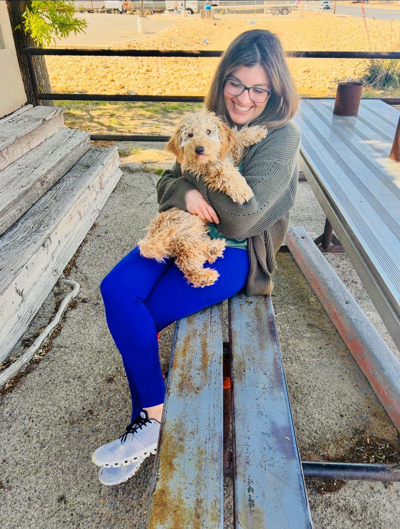 Woman in blue leggings and sweater smiles, holding a tan puppy on a wooden bench outdoors.