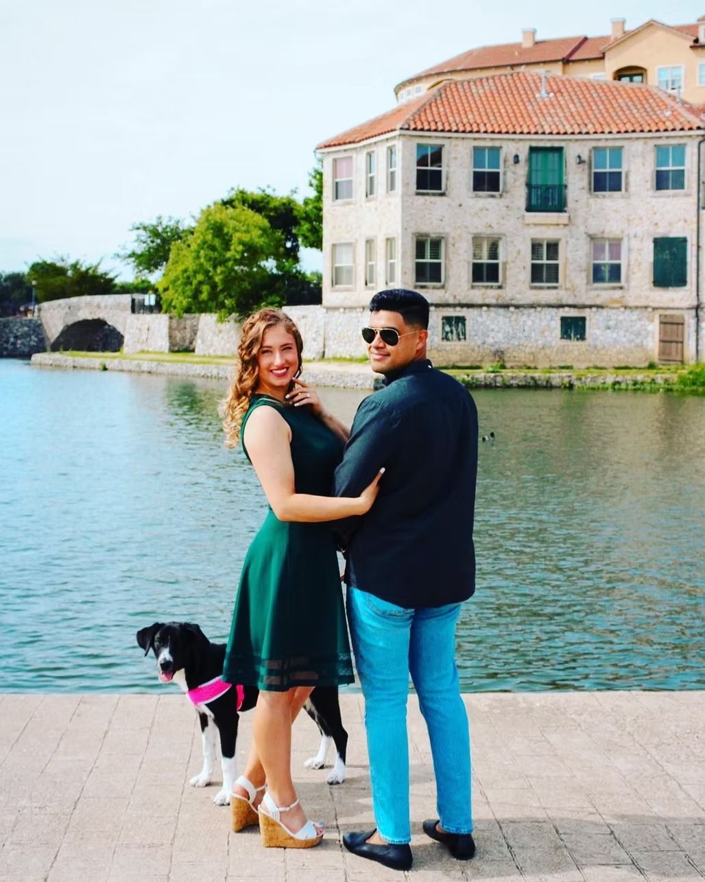 Couple posing by a lake with a dog; the woman in a green dress and heels, the man in a black shirt and jeans, next to a building.