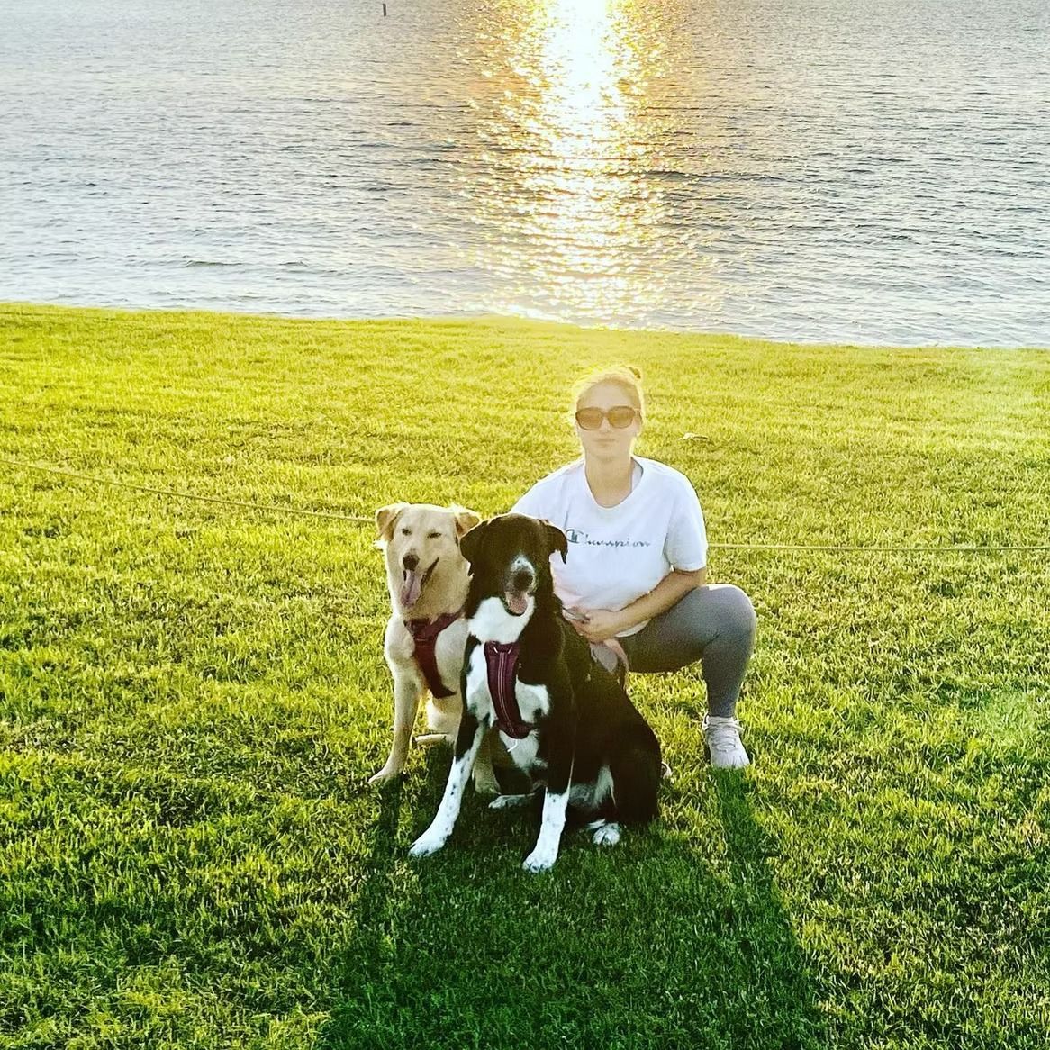 Woman squats with two dogs on a grassy hill near a lake at sunset.