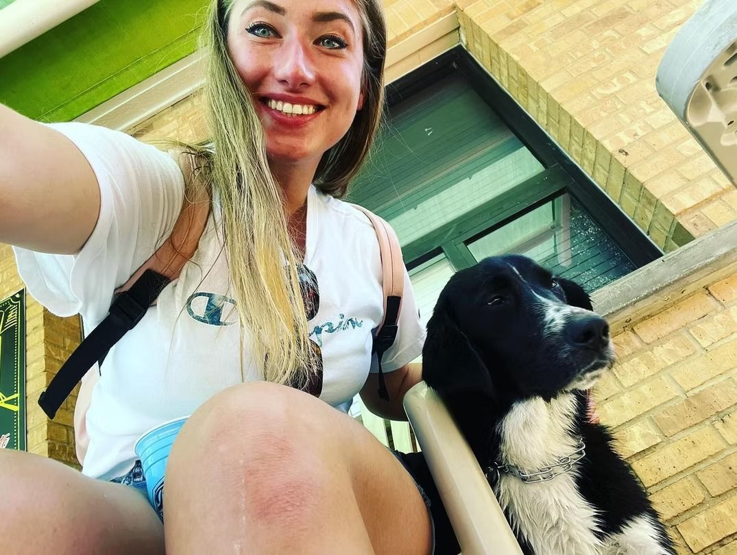 Woman with blonde hair smiles next to a black and white dog outdoors, near a building.