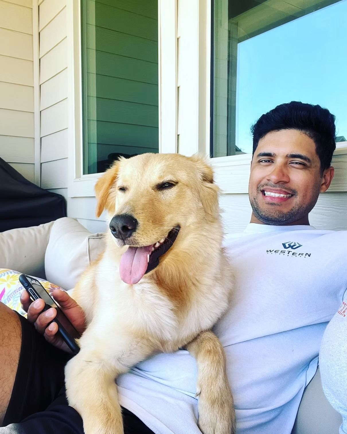 Man smiling with a happy golden dog outdoors on a couch. The dog has its tongue out, both relaxing.
