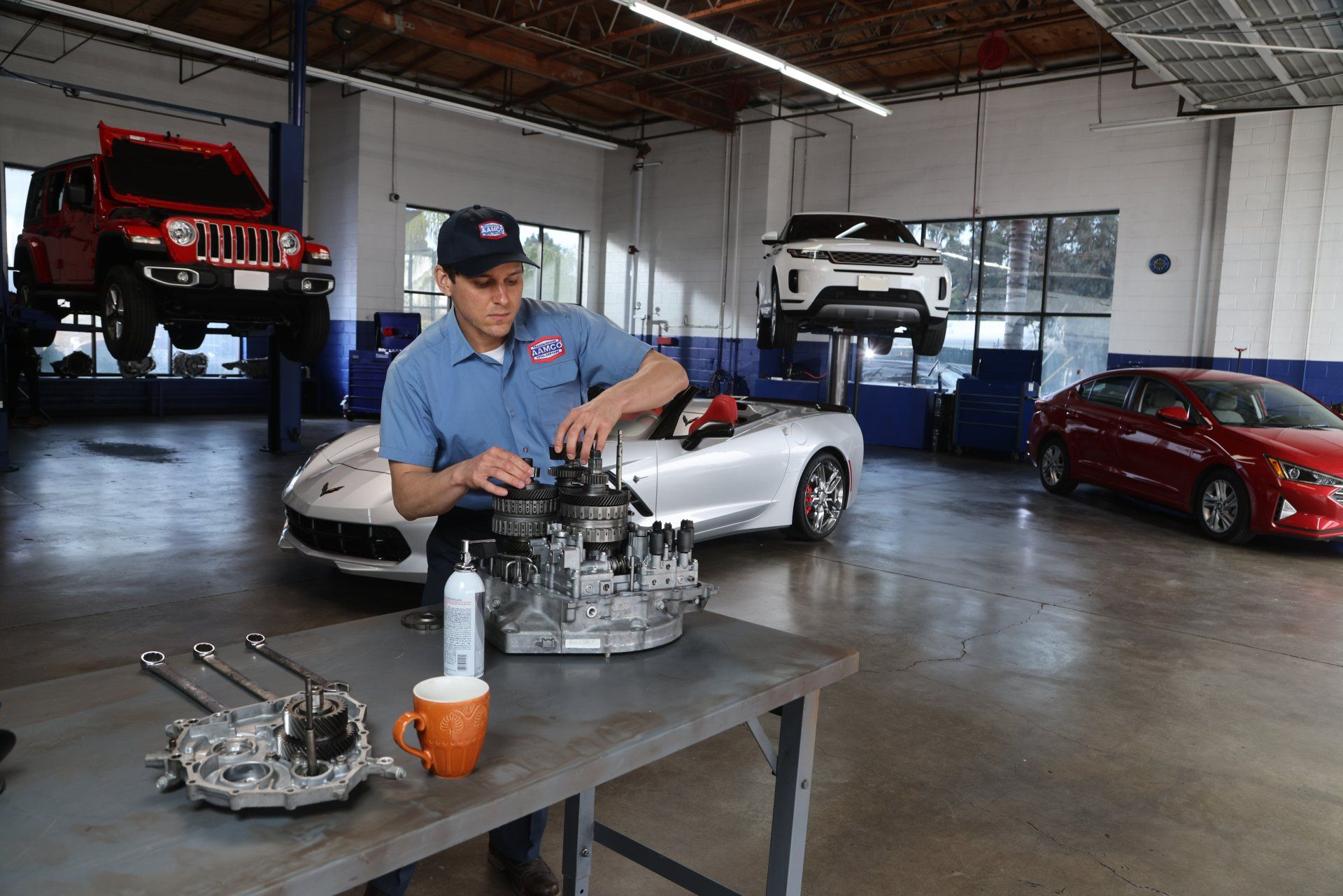 Mechanic working on a car transmission in a repair shop, other vehicles on lifts.