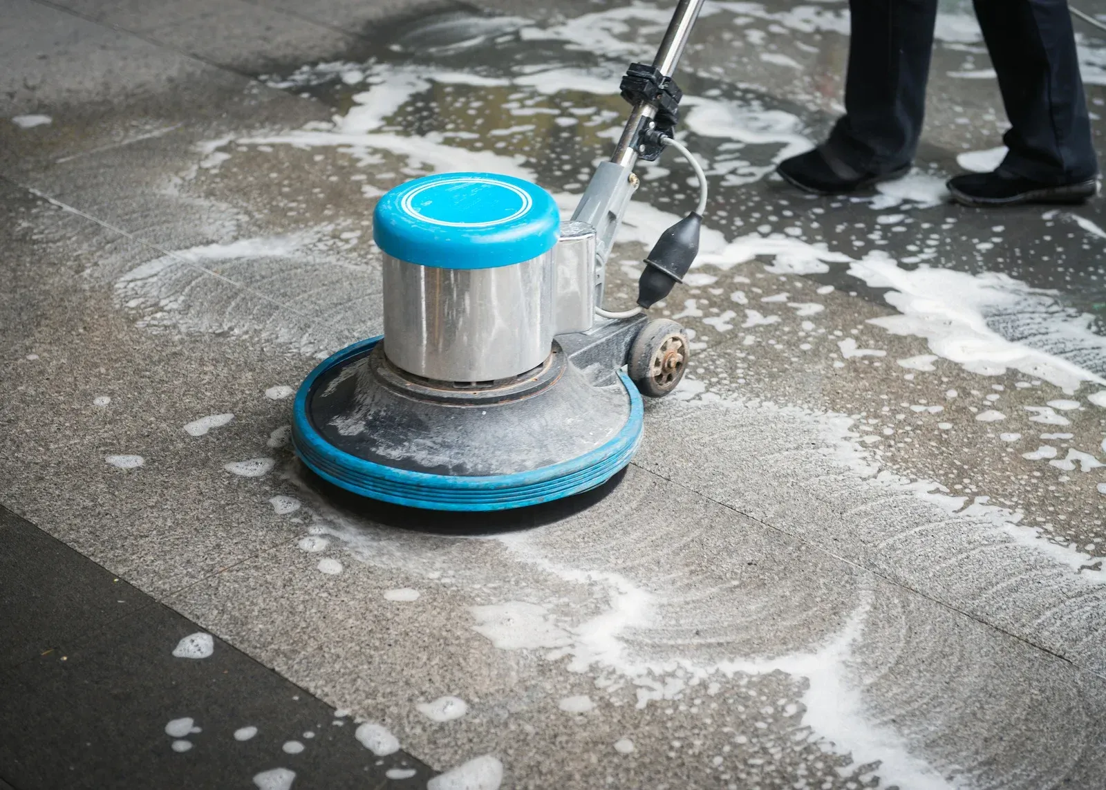 A person using a blue industrial floor scrubber to clean a soapy, tiled floor.