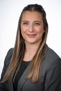 Woman with long blonde hair, wearing a gray blazer and dark green shirt, smiling.