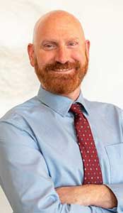 Bald man with red beard and tie, arms crossed, smiles, blue shirt.