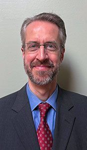 Man in suit and glasses smiling at the camera; red tie, blue shirt, neutral background.