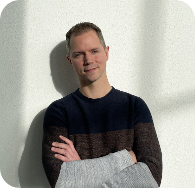 Man in a color-block sweater leaning against a white wall with arms crossed, smiling.