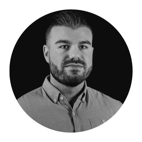 Man with fair skin wearing a dark blue t-shirt, sitting on a gray couch and smiling.