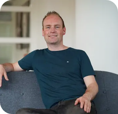 Man with fair skin wearing a dark blue t-shirt, sitting on a gray couch and smiling.